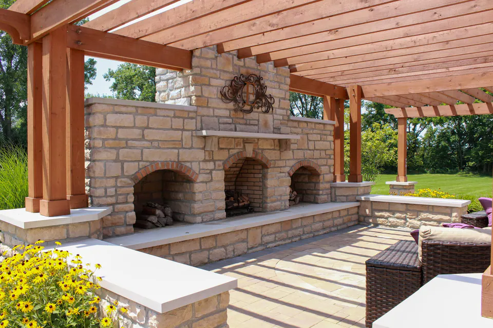 Stone outdoor fireplace beneath wood pergola with paver patio, lounge furniture, and flowering planter.